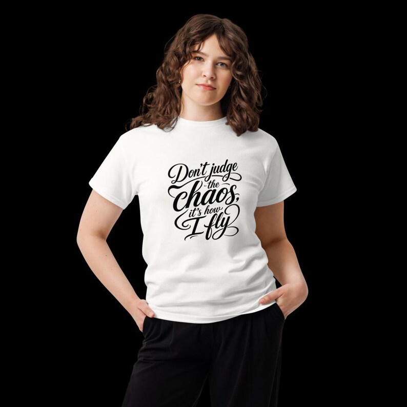 May include: A white short-sleeved t-shirt featuring black cursive text on the front that reads, Don't judge the chaos, it's how I fly. The shirt has a crew neckline and a standard fit, paired with black trousers.