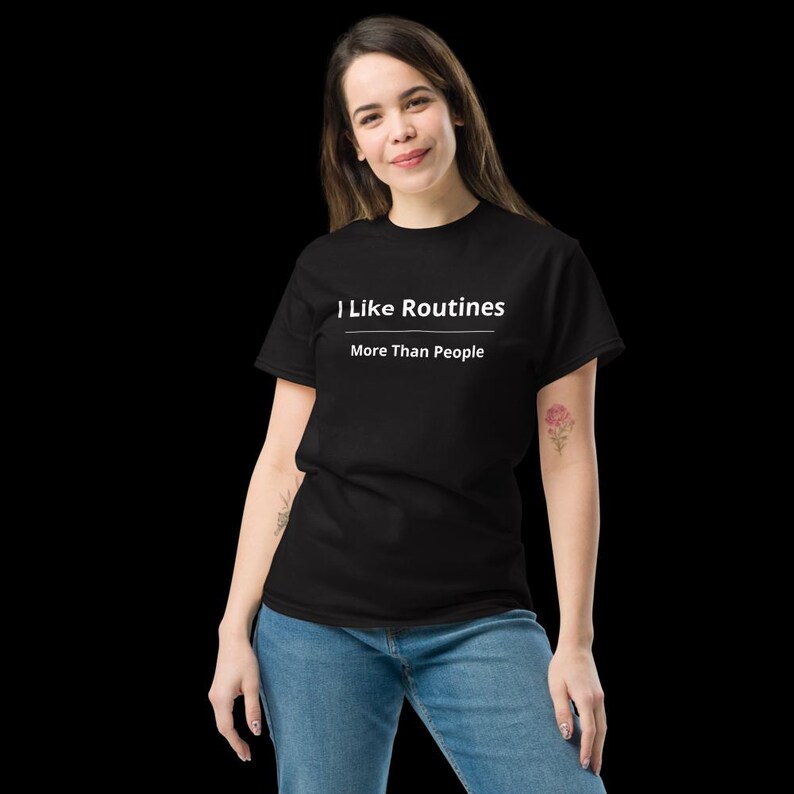 K&ouml;nnte beinhalten: Ein schwarzes kurz&auml;rmeliges T-Shirt mit dem wei&szlig;en Text "I Like Routines" &uuml;ber einer horizontalen Linie und dem Text "More Than People" darunter, kombiniert mit einer blauen Jeans.