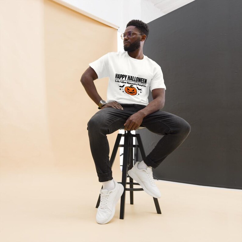 K&ouml;nnte beinhalten: Ein wei&szlig;es kurz&auml;rmeliges T-Shirt mit dem schwarzen Text Happy Halloween To My Fellow Neurospicy Pumpkins &uuml;ber einer Grafik eines orangefarbenen K&uuml;rbisses mit schwarzen Flederm&auml;usen und Sternen, kombiniert mit einer dunkelgrauen Jeans und wei&szlig;en Turnschuhen.