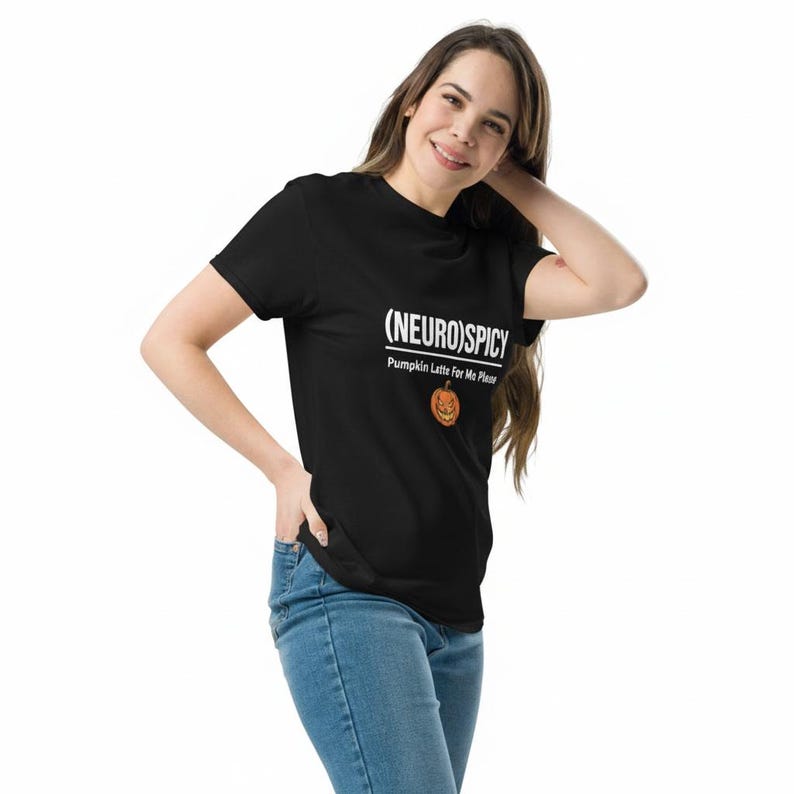 May include: A black short-sleeved t-shirt featuring the white text (NEURO)SPICY and Pumpkin Latte For Me Please, accompanied by a small orange jack-o'-lantern graphic centered on the chest area, paired with blue denim jeans.