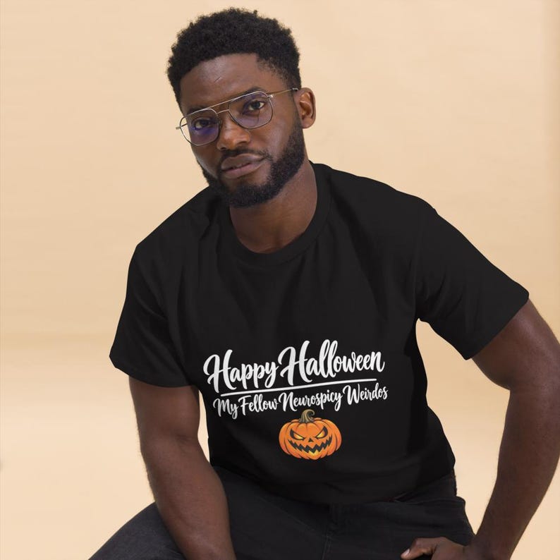 May include: A black short-sleeved t-shirt featuring the white text Happy Halloween My Fellow Neurospicy Weirdos above a graphic of an orange jack-o'-lantern pumpkin with a carved face and glowing yellow eyes.