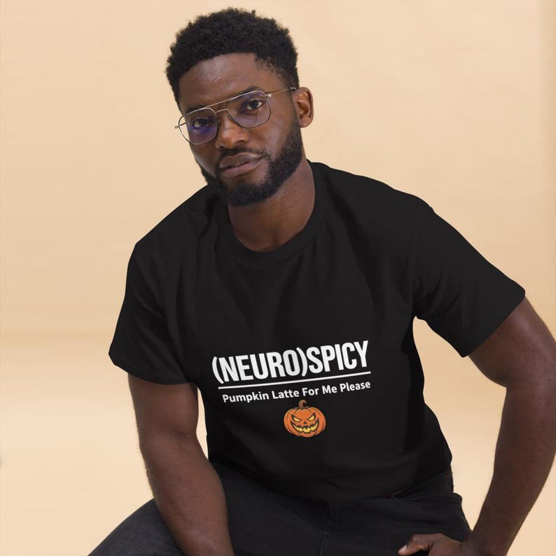 May include: A black short-sleeved t-shirt featuring white text that reads (NEURO)SPICY and Pumpkin Latte For Me Please, accompanied by a small orange jack-o'-lantern graphic centered below the text on the front of the garment.