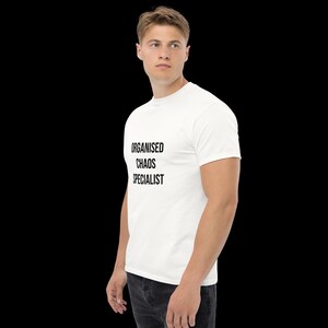 May include: A white short-sleeved t-shirt featuring the black text "ORGANISED CHAOS SPECIALIST" printed on the front. The shirt has a crew neckline and a standard fit, shown from a side profile angle against a plain black background.