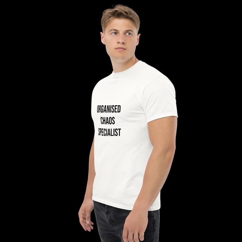 May include: A white short-sleeved t-shirt featuring the black text "ORGANISED CHAOS SPECIALIST" printed on the front. The shirt has a crew neckline and a standard fit, shown from a side profile angle against a plain black background.