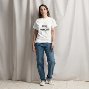 May include: A white short-sleeved t-shirt featuring the black text "CHAOS isn't my hobby, it's my WORKLOAD" printed on the chest, paired with blue denim jeans and white sneakers. The shirt has a crew neckline and a standard fit.