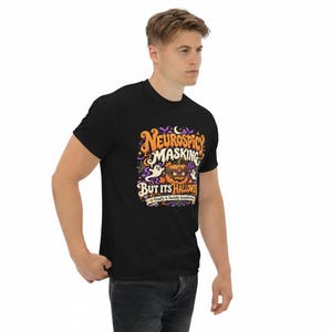 Pu&ograve; includere: Una t-shirt nera con una stampa grafica colorata con il testo "NEUROSPICY MASKING BUT ITS HALLOWEEN" e "Finally A Socially Acceptable Mask". Il design include una zucca con una maschera viola, fantasmi, pipistrelli e stelle.