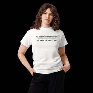 Op de afbeelding: Een wit T-shirt met korte mouwen en de zwarte tekst I Fix The Invisible Disasters, gevolgd door een horizontale lijn en de tekst You Swear You Didn't Cause. Het T-shirt wordt gecombineerd met een zwarte broek voor een eenvoudige uitstraling.