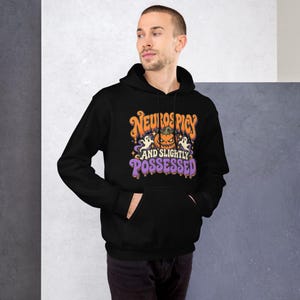 May include: A black hooded sweatshirt featuring a graphic print with the text "NEUROSPICY AND SLIGHTLY POSSESSED" in orange and purple. The design includes a central jack-o'-lantern, two white ghosts, and small yellow stars on the front.