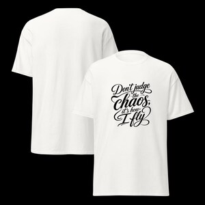 May include: A white short-sleeved t-shirt featuring black cursive text on the front that reads, Don't judge the chaos, it's how I fly. The shirt is displayed from both the front and back angles, showing a plain white back and a graphic front.