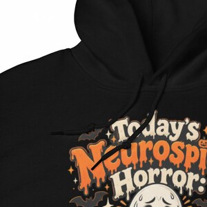 K&ouml;nnte beinhalten: Ein schwarzer Kapuzenpullover mit einem Grafikdruck und dem Text Today's Neurospicy Horror. Das Design zeigt orangefarbene, tropfende Schriftz&uuml;ge, kleine schwarze Flederm&auml;use, Sterne und einen wei&szlig;en Geist mit besorgtem Gesichtsausdruck.