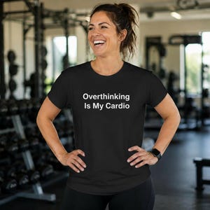 May include: A black short-sleeved t-shirt featuring the white text "Overthinking Is My Cardio" printed across the chest. The garment has a crew neckline and a standard fit, constructed from a solid black fabric with clean, white lettering.