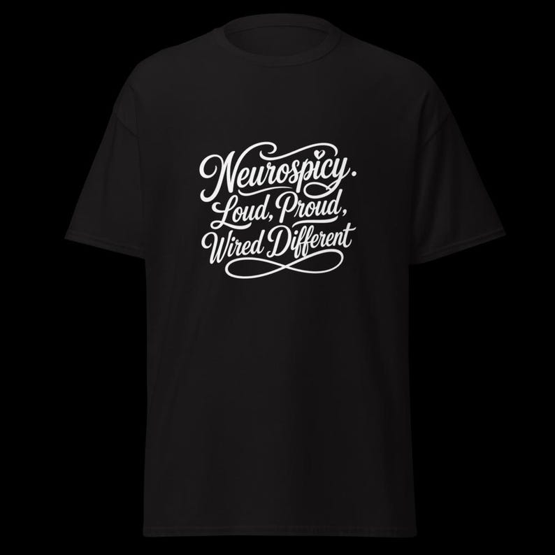 K&ouml;nnte beinhalten: Ein schwarzes Kurzarm-T-Shirt mit wei&szlig;em Schreibschrift-Text auf der Brust, der Neurospicy. Loud, Proud, Wired Different lautet. Das Design enth&auml;lt ein kleines Herz &uuml;ber dem ersten Wort und ein dekoratives Unendlichkeitssymbol unter dem Text.