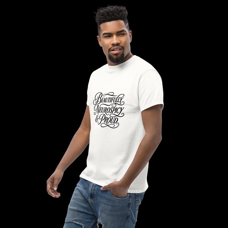 K&ouml;nnte beinhalten: Ein wei&szlig;es Kurzarm-T-Shirt mit schwarzem, kursivem Text auf der Brust, der Beautifully Neurospicy & Proud lautet. Das Shirt hat einen Rundhalsausschnitt und wird mit einer blauen Jeans kombiniert, was ein l&auml;ssiges und klassisches Design ergibt.