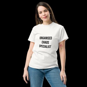May include: A white short-sleeved t-shirt featuring the black text "ORGANISED CHAOS SPECIALIST" printed on the chest. The garment is paired with blue denim jeans, creating a casual look with a clear, bold graphic message on the front of the shirt.