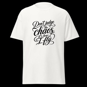 May include: A white short-sleeved t-shirt featuring black text printed on the centre of the chest. The text reads, "Don't judge the chaos, it's how I fly," written in a decorative, flowing script font with varying line weights and flourishes.