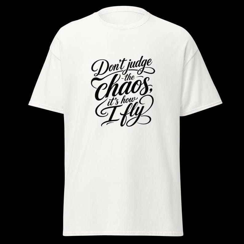 May include: A white short-sleeved t-shirt featuring black text printed on the centre of the chest. The text reads, "Don't judge the chaos, it's how I fly," written in a decorative, flowing script font with varying line weights and flourishes.