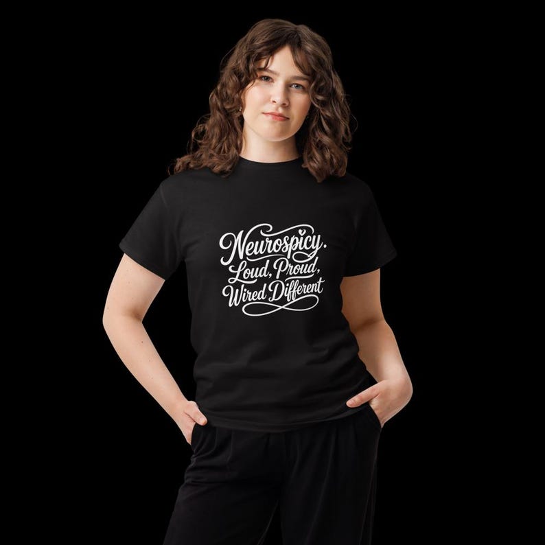 K&ouml;nnte beinhalten: Ein schwarzes Kurzarm-T-Shirt mit wei&szlig;em, kursivem Schriftzug auf der Brust, der Neurospicy. Loud, Proud, Wired Different lautet. Das Oberteil hat einen Rundhalsausschnitt und wird mit einer schwarzen Hose kombiniert, was einen einfarbigen Look ergibt.