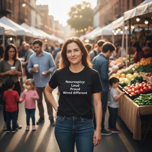 Könnte beinhalten: Ein schwarzes Kurzarm-T-Shirt mit weißem Text, der lautet: Neurospicy: Loud, Proud, Wired different. Das Shirt wird mit einer blauen Jeans kombiniert und zeichnet sich durch einen klassischen Rundhalsausschnitt und eine normale Passform aus.