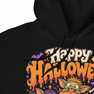 May include: A black hooded sweatshirt featuring a graphic print with the text Happy Halloween in orange and cream dripping letters. The design includes a small purple bat, orange stars, and a green creature with orange hair peeking out.