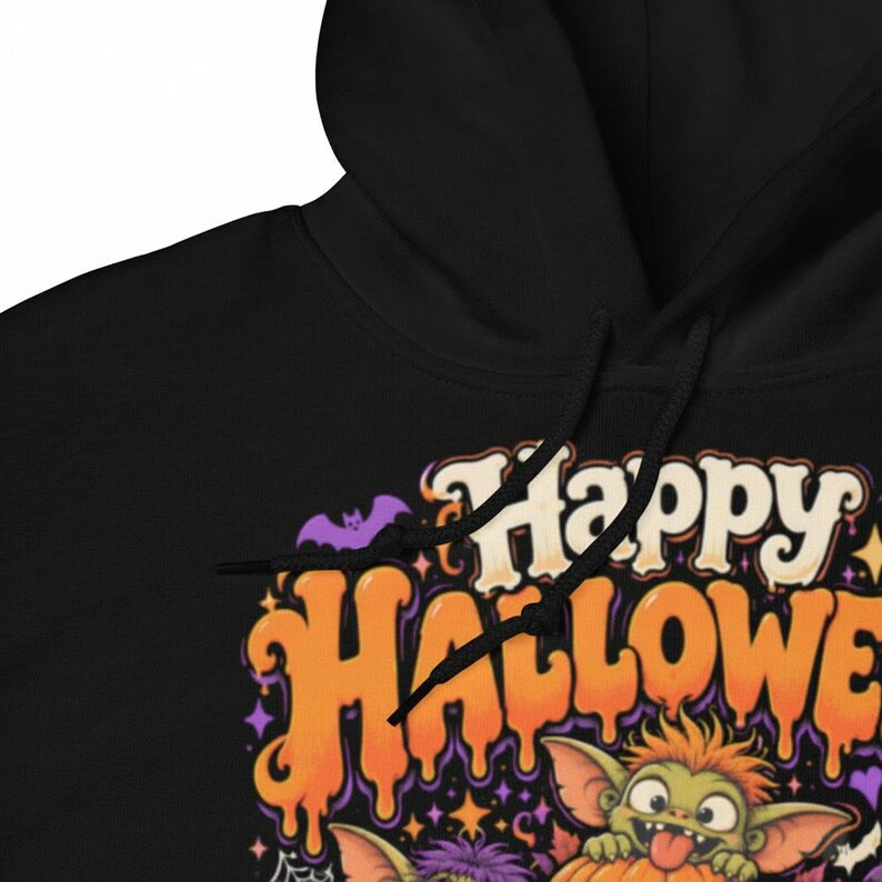 May include: A black hooded sweatshirt featuring a graphic print with the text Happy Halloween in orange and cream dripping letters. The design includes a small purple bat, orange stars, and a green creature with orange hair peeking out.