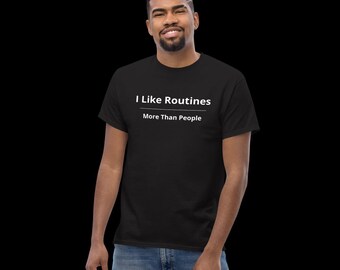 Op de afbeelding: Een zwart t-shirt met korte mouwen en de witte tekst I Like Routines More Than People op de borst gedrukt. Het ontwerp bevat een horizontale witte lijn die de twee zinnen scheidt, gecentreerd op de voorzijde van het kledingstuk.