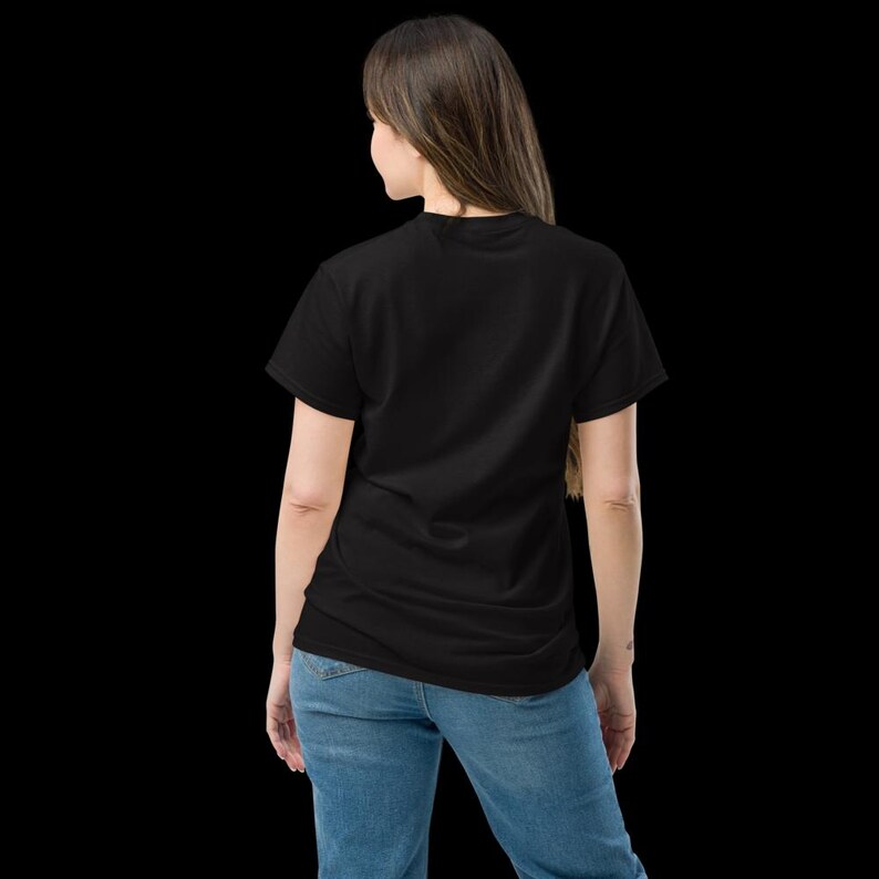 K&ouml;nnte beinhalten: Ein schlichtes schwarzes T-Shirt mit kurzen &Auml;rmeln und Rundhalsausschnitt, kombiniert mit einer blauen Jeans. Das T-Shirt ist durchgehend schwarz gef&auml;rbt, was f&uuml;r ein schlichtes und neutrales Erscheinungsbild in Kombination mit der blauen Hose sorgt.