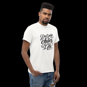 May include: A white short-sleeved t-shirt featuring black cursive text on the chest that reads, Don't judge the chaos, it's how I fly. The shirt has a crew neckline and is paired with blue denim jeans that have a visible distressed detail.