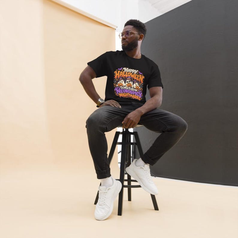 May include: A black short-sleeved t-shirt featuring a colourful graphic print with the text Happy Halloween To My Fellow Neurospicy Nightmares, depicting three cartoon ghosts wearing hats and sunglasses surrounded by pumpkins and bats.