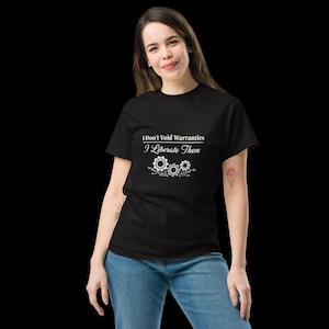 Op de afbeelding: Een zwart t-shirt met korte mouwen en witte tekst die luidt I Don't Void Warranties I Liberate Them, vergezeld van een afbeelding van drie onderling verbonden mechanische tandwielen en printplaatlijnen op de borst.
