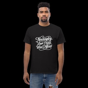 K&ouml;nnte beinhalten: Ein schwarzes kurz&auml;rmeliges T-Shirt mit wei&szlig;em Schreibschrift-Text auf der Brust, der Neurospicy. Loud, Proud, Wired Different lautet. Das T-Shirt hat einen Rundhalsausschnitt und wird mit einer blauen Jeans kombiniert, die am Knie abgenutzt ist.