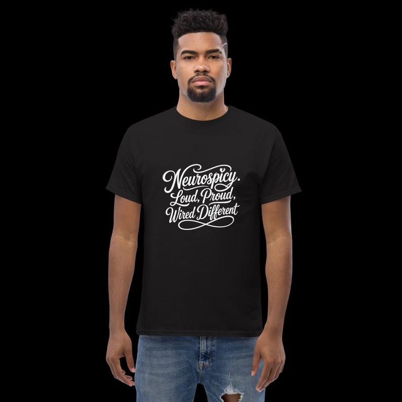 K&ouml;nnte beinhalten: Ein schwarzes kurz&auml;rmeliges T-Shirt mit wei&szlig;em Schreibschrift-Text auf der Brust, der Neurospicy. Loud, Proud, Wired Different lautet. Das T-Shirt hat einen Rundhalsausschnitt und wird mit einer blauen Jeans kombiniert, die am Knie abgenutzt ist.