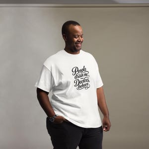 Op de afbeelding: Een wit t-shirt met korte mouwen en zwarte cursieve tekst op de borst met de tekst People drain me Devices behave. Het shirt heeft een ronde hals en een standaard pasvorm, gecombineerd met een zwarte broek en een horloge om de linkerpols.