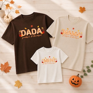 May include: A dark brown t-shirt with the text DADA, a beige t-shirt with the text MAMA, and a white t-shirt with the text MINI. Each shirt features a plaid pattern within the letters, autumn leaf graphics, and the phrase grateful & thankful.