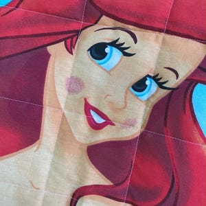 May include: A close-up of a quilted fabric featuring a cartoon illustration of a character with red hair, blue eyes, and a purple top. The character's face is centered, with a light skin tone and a smiling expression. The background is a light blue colour.