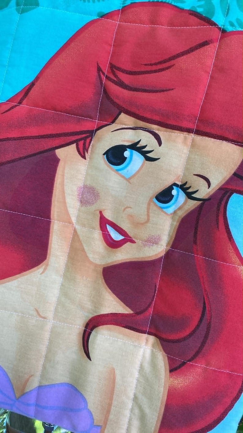 May include: A close-up of a quilted fabric featuring a cartoon illustration of a character with red hair, blue eyes, and a purple top. The character's face is centered, with a light skin tone and a smiling expression. The background is a light blue colour.