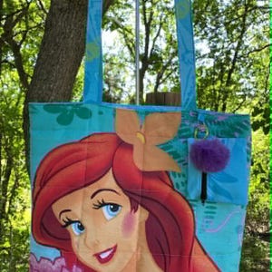 May include: A teal tote bag featuring a colourful illustration of Ariel from The Little Mermaid. The bag has teal straps and a small pocket with a purple pom-pom. The background is a lush green.