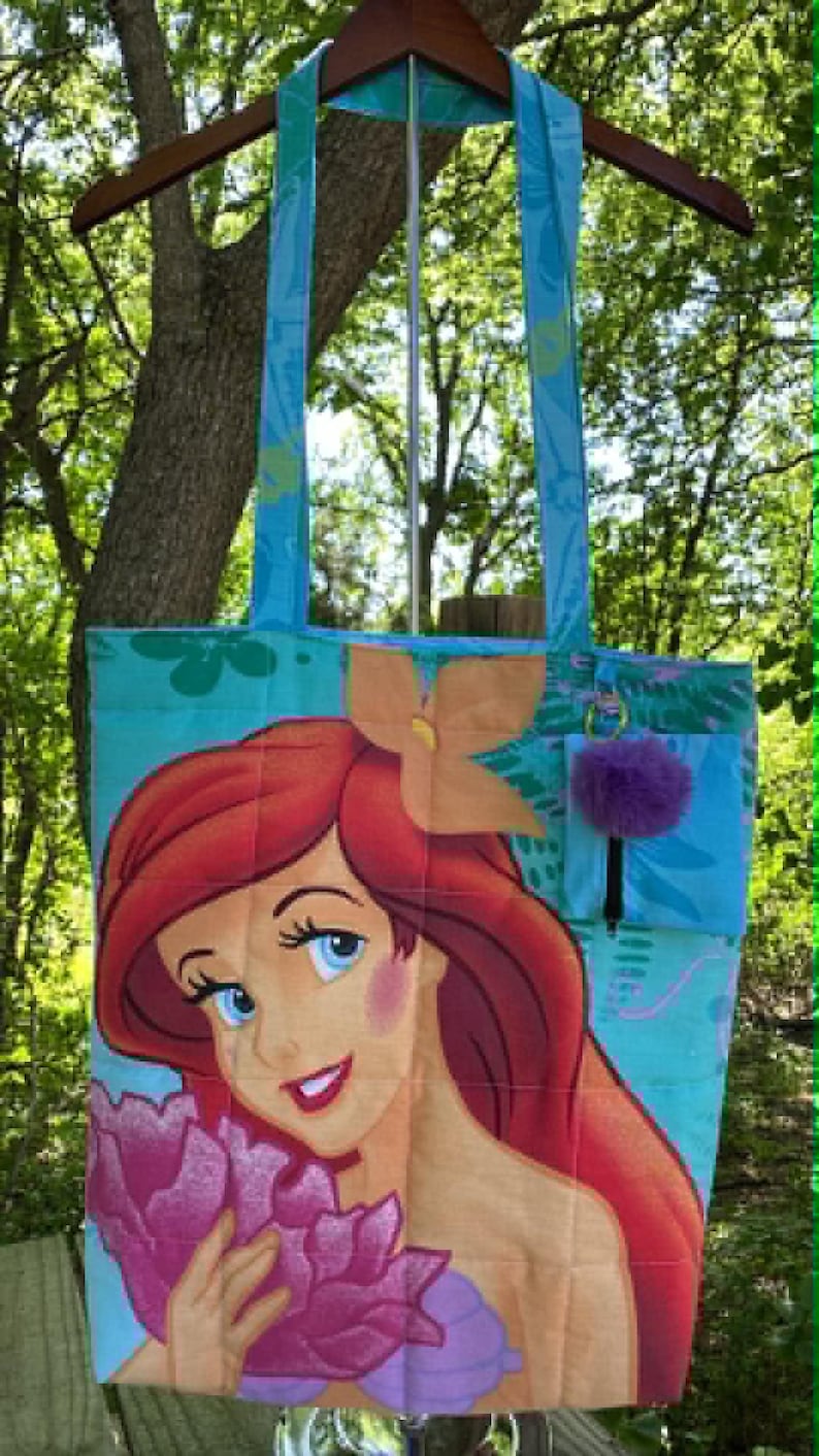May include: A teal tote bag featuring a colourful illustration of Ariel from The Little Mermaid. The bag has teal straps and a small pocket with a purple pom-pom. The background is a lush green.