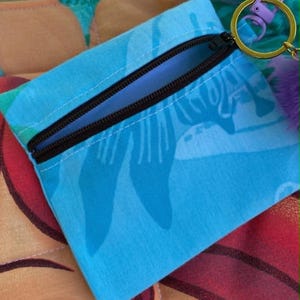 May include: A small, blue zippered pouch with a black zip and a key ring. The pouch features a tropical pattern in shades of blue and white. A purple pom-pom is attached to the key ring. The pouch is on top of a larger bag with a similar pattern.