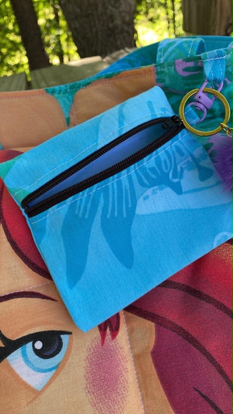 May include: A small, blue zippered pouch with a black zip and a key ring. The pouch features a tropical pattern in shades of blue and white. A purple pom-pom is attached to the key ring. The pouch is on top of a larger bag with a similar pattern.