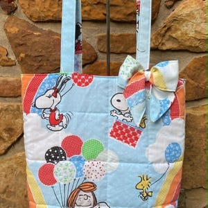 May include: Quilted tote bag featuring Snoopy and Woodstock characters with balloons and rainbows on a light blue background. The bag has a matching bow and handles, perfect for carrying books or groceries.