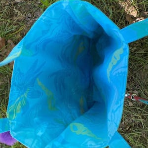 May include: A turquoise tote bag with a floral pattern in shades of blue and green. The bag has matching handles and a purple pom-pom accent. The interior of the bag is visible, showcasing the same patterned fabric.