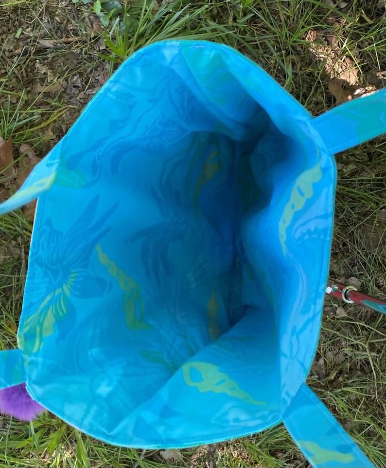 May include: A turquoise tote bag with a floral pattern in shades of blue and green. The bag has matching handles and a purple pom-pom accent. The interior of the bag is visible, showcasing the same patterned fabric.