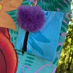May include: A colourful tote bag with a floral pattern in shades of orange, red, and teal. It features a small blue pocket with a black zip and a purple pom-pom attached with a keychain. The bag is outdoors.