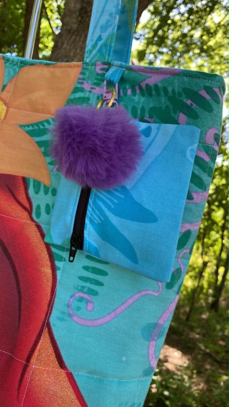 May include: A colourful tote bag with a floral pattern in shades of orange, red, and teal. It features a small blue pocket with a black zip and a purple pom-pom attached with a keychain. The bag is outdoors.
