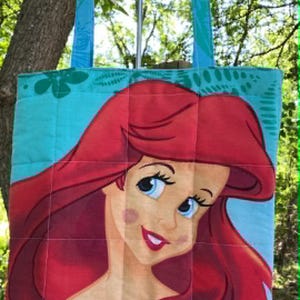 May include: A quilted tote bag with a large illustration of a red-haired mermaid with blue eyes and a purple top. The bag has teal handles with a leaf pattern and a teal background.