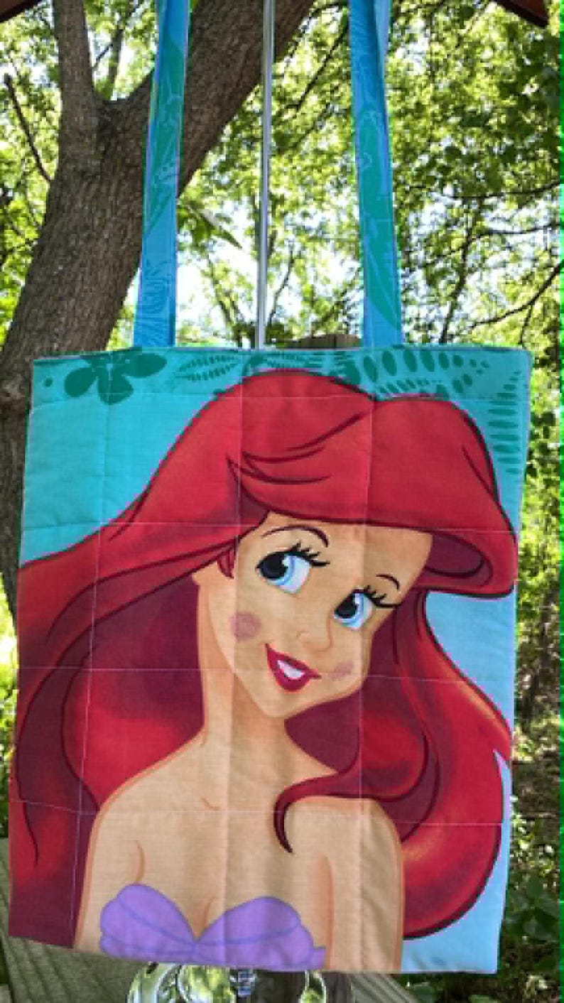 May include: A quilted tote bag with a large illustration of a red-haired mermaid with blue eyes and a purple top. The bag has teal handles with a leaf pattern and a teal background.