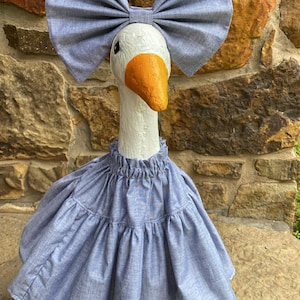May include: A decorative goose statue with a white head, orange beak, and a blue bow and dress. The goose is standing on a green base, and the background is a stone wall.