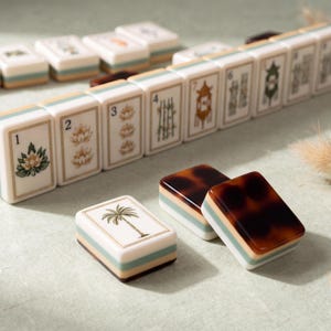 May include: A set of rectangular mahjong tiles featuring cream-colored faces with various illustrations including numbered lotus flowers, bamboo, and a palm tree. The tiles have a layered design with a light blue stripe and a dark brown tortoiseshell back.
