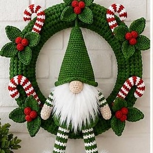 Op de afbeelding: Een gehaakte groene krans met een centrale kabouter met een hoge groene muts, een witte imitatiebont baard en gestreepte benen. De krans is versierd met rood-witte zuurstokken en clusters van groene bladeren met kleine rode bessen.