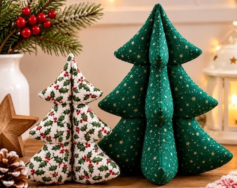 Op de afbeelding: Twee stoffen kerstboomornamenten. Eén boom is wit met een patroon van groene hulstbladeren en rode bessen. De tweede boom is donkergroen met een gouden sterrenprint. Beide bomen hebben een zachte, driedimensionale, gelaagde constructie.